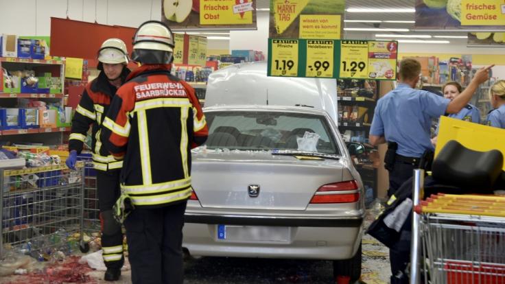 Feuerwehrleute und Polizeibeamte stehen in einem Supermarkt neben einem Pkw, der zuvor in den Supermarkt gefahren war und dabei fünf Menschen verletzt hat.