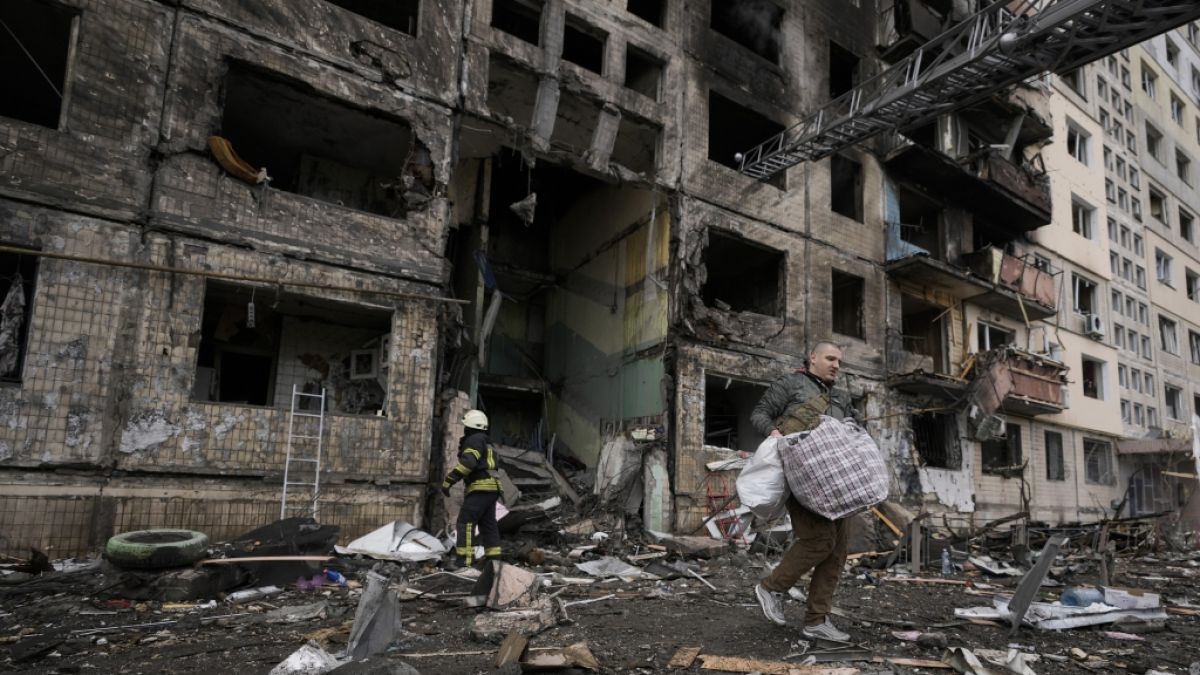 Ein Mann verlässt mit einer Tasche ein zerstörtes Wohngebäude, vor dem ein Feuerwehrmann im Einsatz ist. (Foto)
