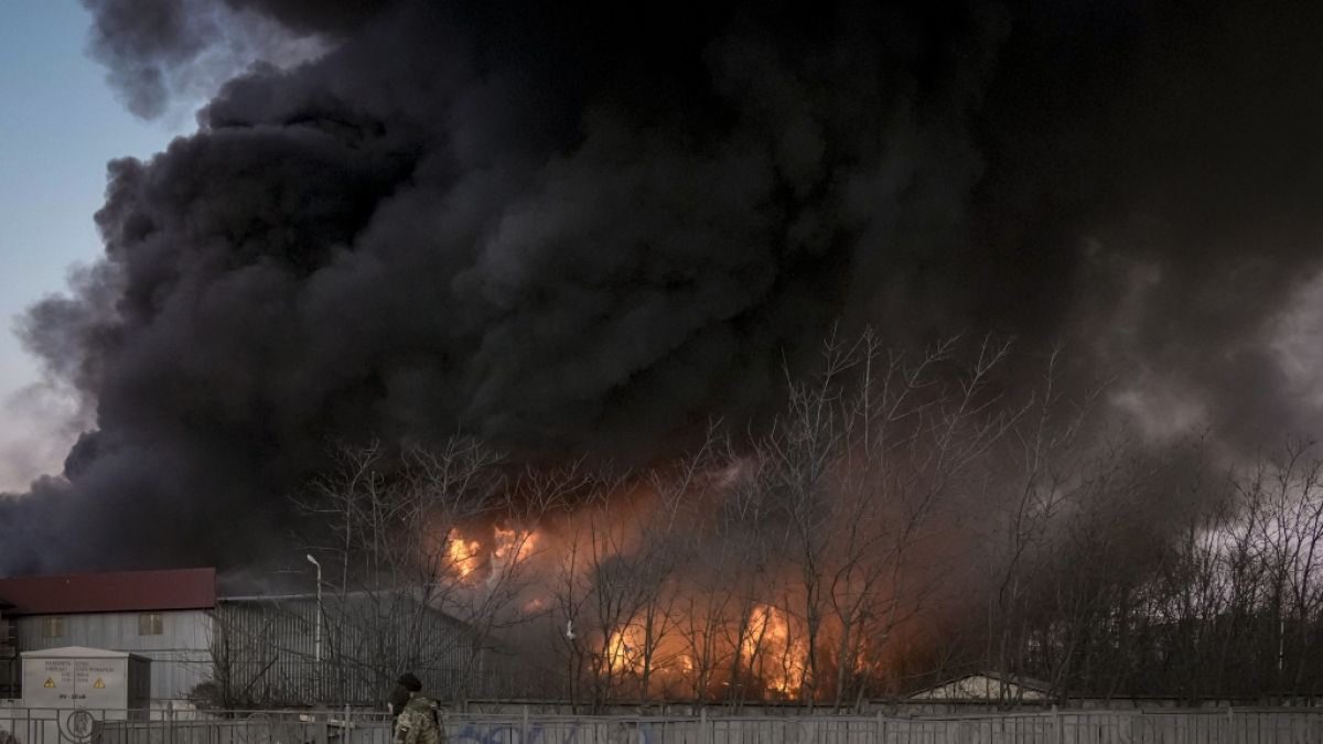 Ukrainische Soldaten tragen einen Container während im Hintergrund ein Lagerhaus nach einem Bombenanschlag in Flammen steht. (Foto)