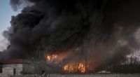 Ukrainische Soldaten tragen einen Container während im Hintergrund ein Lagerhaus nach einem Bombenanschlag in Flammen steht.