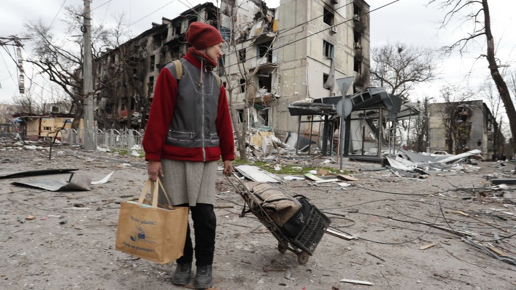 Eine Anwohnerin geht am 18.04.2022 mit Gepäck an einem beschädigten Wohnhaus vorbei. Die Hafenstadt Mariupol ist seit dem 1. März 2022 vollständig von russischen Truppen eingeschlossen und mittlerweile wohl komplett erobert. (Foto)