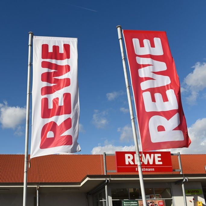 Fahnen mit dem REWE-Logo wehen vor einer Filiale des Einzelhändlers (Symbolbild).