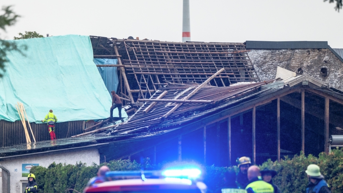 Einsatzkr&auml;fte sichern das zerst&ouml;rte Dach eines Hauses mit Planen gegen das Wetter w&auml;hrend Feuerwehrleute sich vor einem zerst&ouml;rten Haus beraten. (Foto)