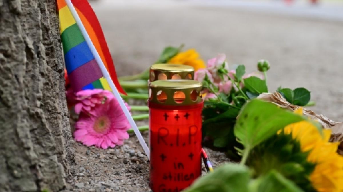 Eine Regenbogenflagge, Blumen und Kerzen stehen vor einem Baum nahe dem Bereich, wo ein Fahrzeug einen Baum gerammt hatte. (Foto)